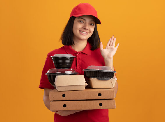 拿着身穿蓝色制服 头戴鸭舌帽的年轻送货女孩 手举比萨饼盒和食品包 微笑着快乐而积极地站在橙色的墙上制服包装盒子
