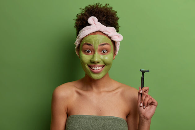 剃刀积极的卷发女人拿着剃须刀 去刮腿毛 脸上敷保湿面膜 关心自己 裹着浴巾 隔离在绿色的墙壁上幸福 纯洁 卫生洗澡卷发新鲜