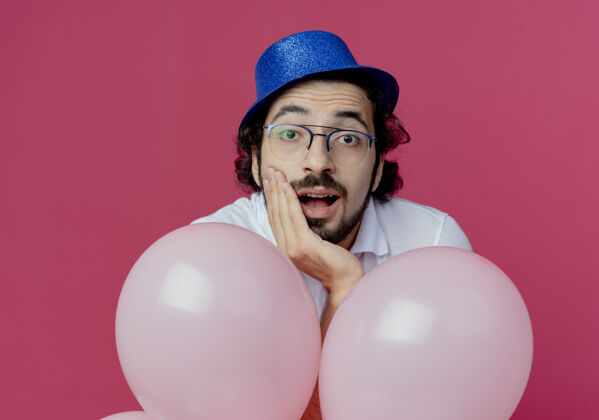 帅哥高兴的帅哥戴着眼镜 戴着蓝色帽子 手里拿着气球 把手放在下巴上 隔离在粉红色的背景上气球帽子拜托