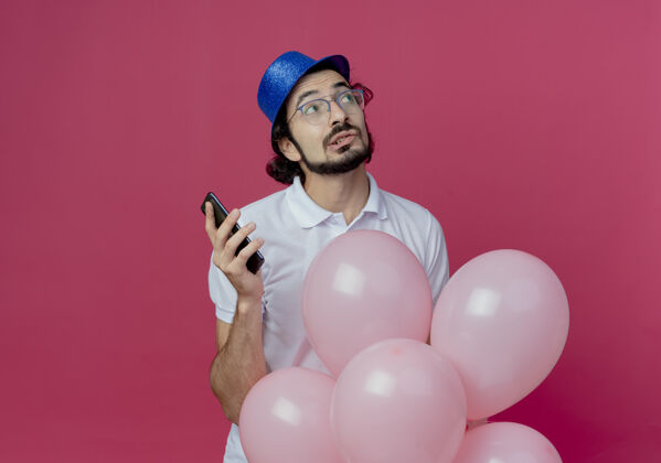 穿看着身边想着帅哥戴着眼镜和蓝色帽子拿着气球和手机孤立在粉色背景上举行壁板眼镜