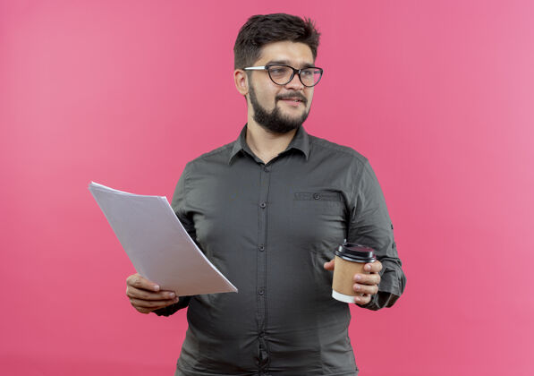手持看着旁边高兴的年轻商人戴着眼镜拿着纸和咖啡杯隔离在粉红色的背景上壁板年轻人咖啡