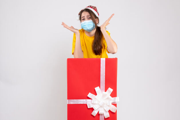 大前视图困惑的女孩 戴着圣诞帽和医用面具站在大圣诞礼物后面礼品立场帽子