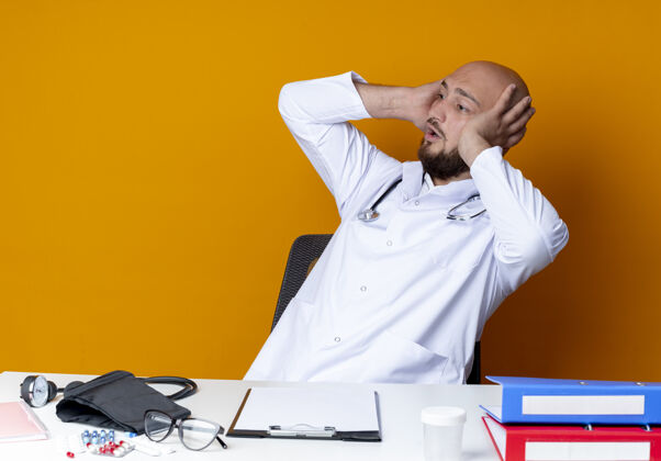 秃头看着一旁惊讶的年轻秃头男医生穿着医用长袍和听诊器坐在办公桌前 拿着医疗工具把脑袋孤立在橙色的背景上头工具壁板