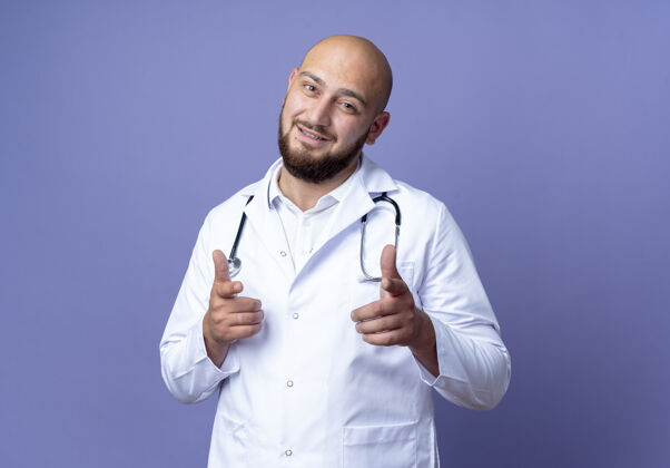 医疗高兴的年轻秃头男医生穿着医用长袍和听诊器显示您的姿态孤立的蓝色背景穿手势表演