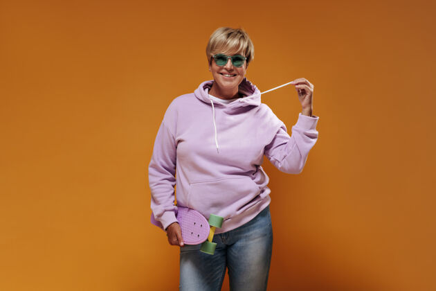 连帽衫一位微笑的女士 短发 戴着现代眼镜 穿着粉色运动衫和酷牛仔裤 在橙色的背景下摆出现代滑板的姿势老年人成熟50多岁