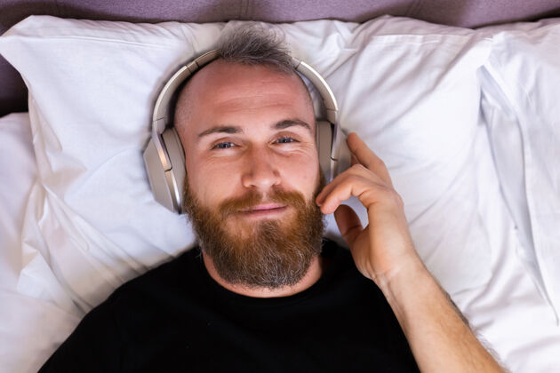 周末快乐的白人男子戴着耳机躺在床上听他最喜欢的音乐 独自休息 跳舞休息耳机房子