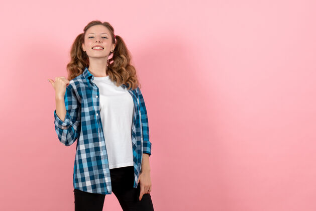 姿势正面图身着蓝色格子衬衫的年轻女性在粉色背景上摆姿势儿童青春情感模特时尚女性色彩人年轻衬衫