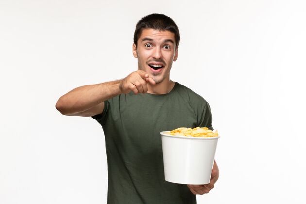 视图正面图身穿绿色t恤的年轻男性手持土豆cips 在浅白的墙上看电影孤独的电影电影院杯子早餐
