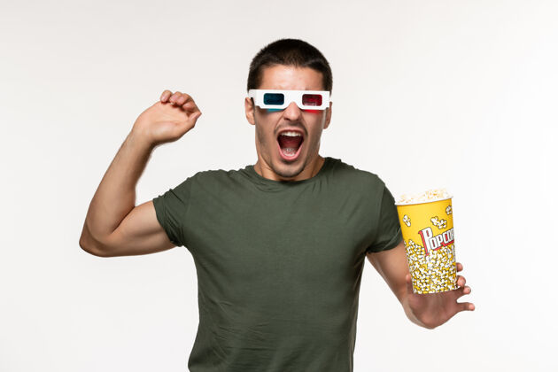 成人正面图身穿绿色t恤的年轻男子手持爆米花 戴着d型太阳镜 在一个白色的电影《孤独的电影院》上表演电影电影穿
