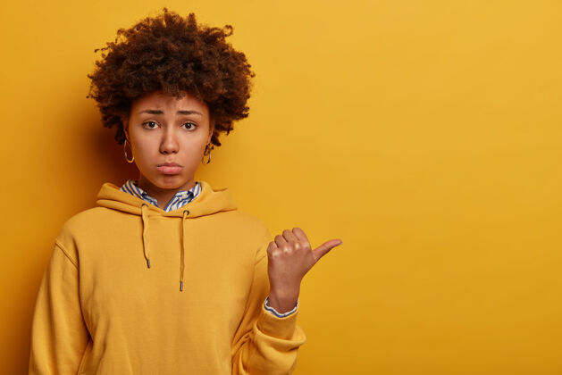 负面不满意的黑皮肤女人的肖像用拇指指向别处 给出负面反馈 对令人失望的事情做出反应 抱怨和抱怨 穿着休闲连帽衫 隔离在黄色的墙上反应情绪建议