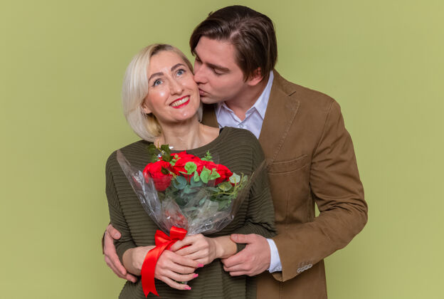 年轻年轻美丽的夫妇幸福的男人和女人捧着一束红玫瑰拥抱着幸福的爱情一起庆祝国际妇女节站在绿色的墙上花束玫瑰情侣