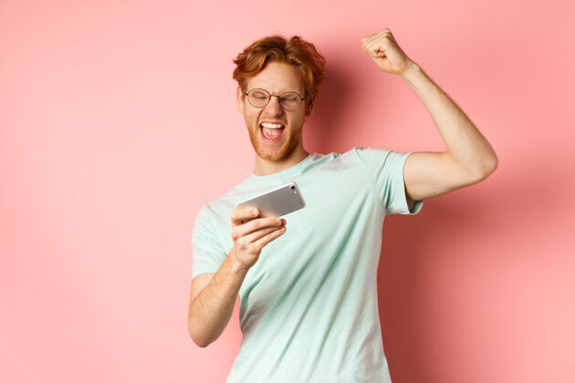 姜快乐的红发人在手机视频游戏中获胜 举起手 高兴地喊“是” 庆祝胜利 看着智能手机 站在粉色背景上男T恤人