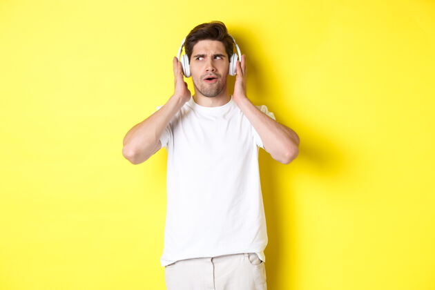 时尚好奇的家伙戴着耳机欣赏音乐 戴着耳机近距离聆听音乐 站在黄色背景上工作室表情男人