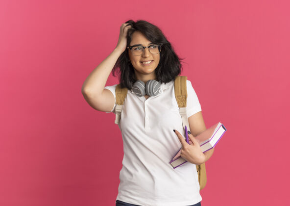 女学生年轻笑容可掬的白人女学生 脖子上戴着耳机 戴着眼镜 背着书包 手放在头发上 手里拿着书 看着粉色的侧面和复印空间背部穿抱着