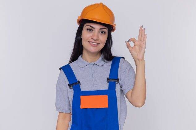 积极身穿施工制服 头戴安全帽的年轻建筑女工人 面带微笑 站在白色的墙上 脸上挂着“好”字年轻展示站着