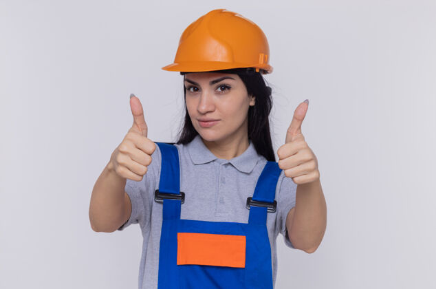 站立身穿建筑制服 头戴安全帽的年轻建筑女工人站在白墙上 面带微笑 自信 快乐 热情地竖起大拇指建筑快乐展示