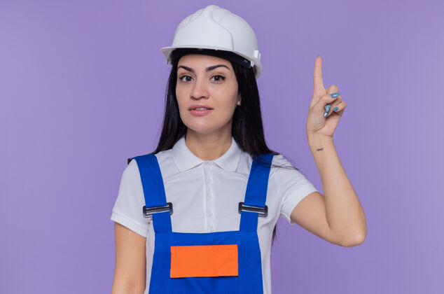 显示身穿施工制服 头戴安全帽的年轻建筑工人站在紫色的墙上 面带微笑地看着前方 露出食指 有着伟大的新想法目录手指建筑