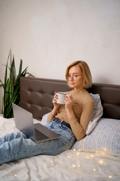 电脑女人在卧室里用笔记本电脑放松 喝一杯热咖啡或茶卧室茶咖啡
