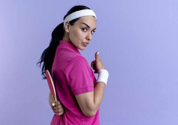 紫色年轻自信的高加索运动女性戴着头带和腕带站在一边拿着球拍在后面和拇指向上紫色的复制空间向上女人球拍