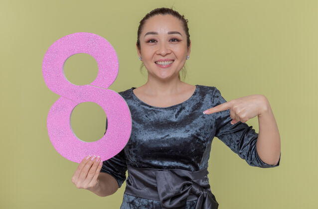 手指快乐的亚洲女人拿着纸板做的数字8 用食指指着它微笑着站在绿色的墙上欢庆国际妇女节举行纸板欢呼