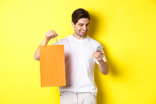 帅气概念折扣 网上银行和现金返还快乐的家伙展示购物袋 看着满意的手机屏幕 黄色背景模特工作室男人