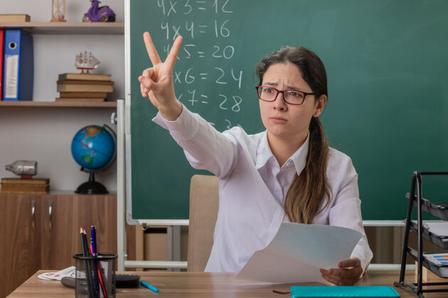 教室戴眼镜的年轻女教师坐在课桌旁 拿着白页检查作业 教室里的黑板前 第二个手指不高兴坐着页面穿
