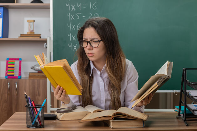 眼镜年轻的女老师戴着眼镜拿着书 坐在教室黑板前的课桌旁 神情迷茫 十分焦急坐着前面非常