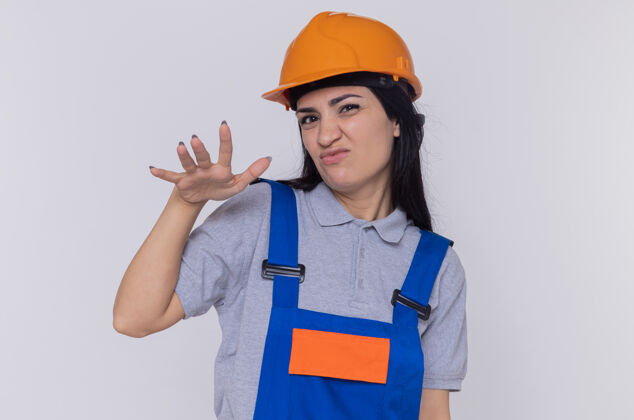 头盔身穿施工制服 头戴安全帽的年轻建筑工人站在白墙上 用手做手势 脸上带着怀疑的表情女人怀疑手势