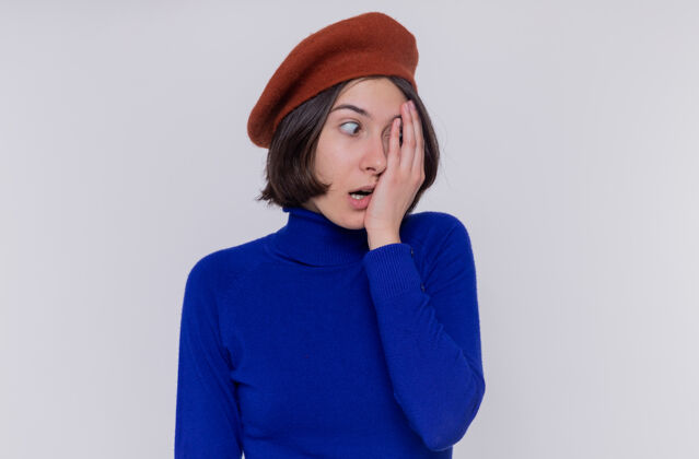 高领毛衣身穿蓝色高领毛衣 头戴贝雷帽 留着短发的年轻女子一边看着一边 一边用手捂住一只眼睛 一边站在白色的墙上女人眼睛盖