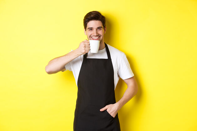 成年人咖啡厅的咖啡师一边喝着咖啡 一边微笑着 穿着黑色围裙 站在黄色的背景上自信烹饪厨师