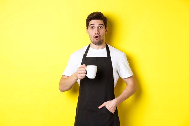 自信咖啡师拿着咖啡杯 看起来很惊讶 穿着黑色围裙咖啡制服站在黄色背景下模特时尚工作室