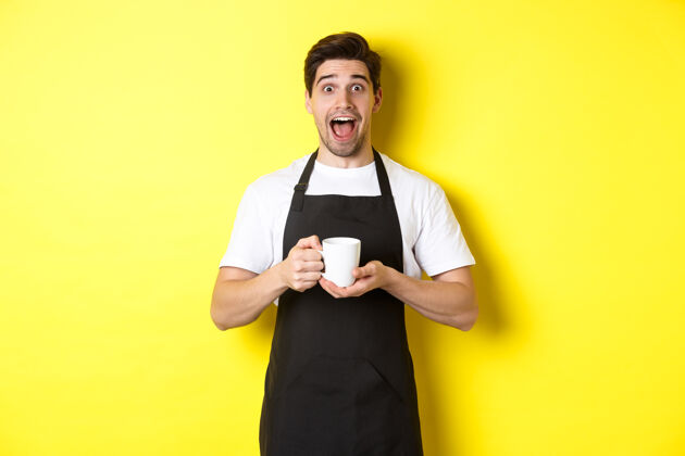年轻友好的咖啡店服务员举手站立 放置您的标志或徽标 站在黄色背景上烹饪帅哥男人