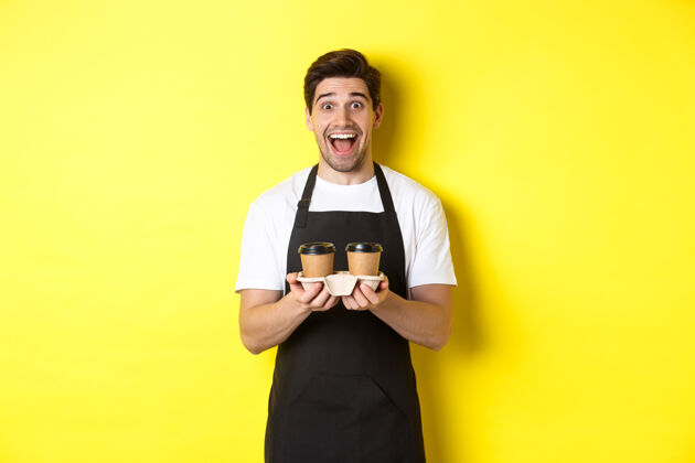 围裙兴奋的男咖啡师拿着两杯外卖咖啡 在咖啡馆工作 站在黄色的背景上自信男士咖啡