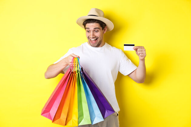 成人度假和理财的概念快乐男人购物者看着购物袋满意 出示信用卡 站在黄色背景下男人工作室黄色