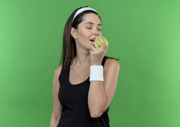 咬戴着头巾的年轻健身女士手拿绿苹果咬着它快乐而积极地站在绿色的墙上苹果年轻站着
