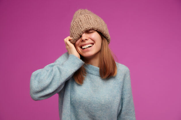 面部迷人的年轻女子的肖像 深色长发 穿着蓝色毛衣 戴着针织帽子 戴着帽子遮住眼睛 在紫色的墙上微笑心情休闲微笑
