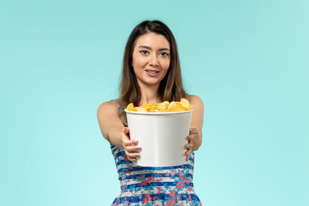杯子正面图：年轻的女性拿着一包薯片 微笑着站在蓝色的表面上电影容器前面
