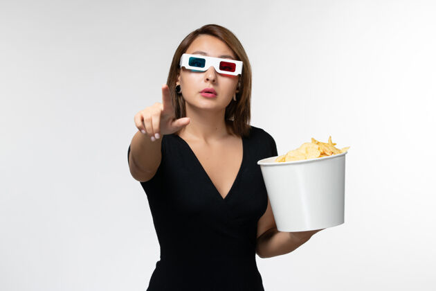 漂亮正面图年轻女性戴着d型太阳镜拿着薯片 在浅白的表面上看电影年轻女性抱着太阳镜