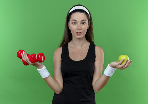 头带戴着头戴耳机的年轻健身女士举着哑铃和绿苹果站在绿色的墙上苹果女人抱着