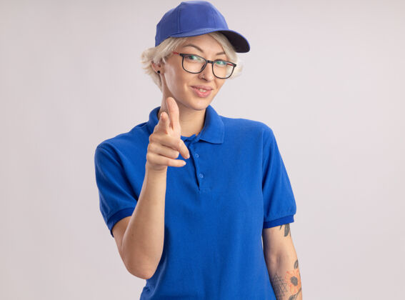 目录身穿蓝色制服 戴着帽子 食指微笑 自信地站在白色墙壁上的年轻女送货员自信手指制服