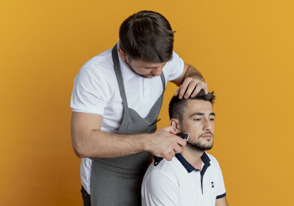 男人穿着围裙的理发师用满意客户的胡须修剪器在橘色墙上刮胡子满意胡须顾客