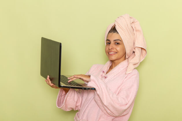 漂亮前视图穿着粉红色浴袍的年轻女性在绿色表面上使用笔记本电脑衣服自我照顾年轻女性