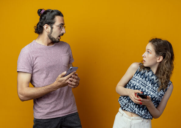 年轻年轻漂亮的夫妇站在橘红色的墙上 拿着智能手机 表情困惑地看着对方站着夫妻每一个