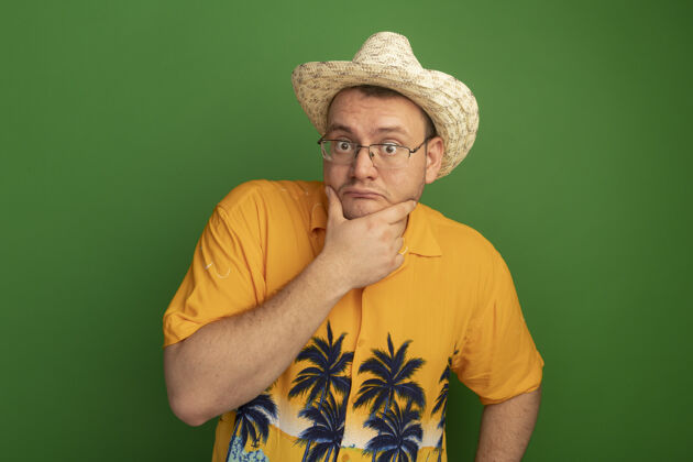 夏天戴着橘色衬衫和夏帽的戴眼镜的男人站在绿色的墙壁上困惑不解站穿男人