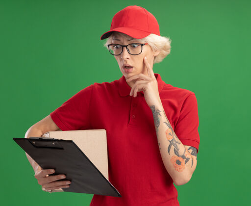 穿身穿红色制服 头戴帽子 戴着眼镜 手拿纸板箱的年轻送货员站在绿色的墙上 神情困惑地看着手中的剪贴板表情剪贴板立场