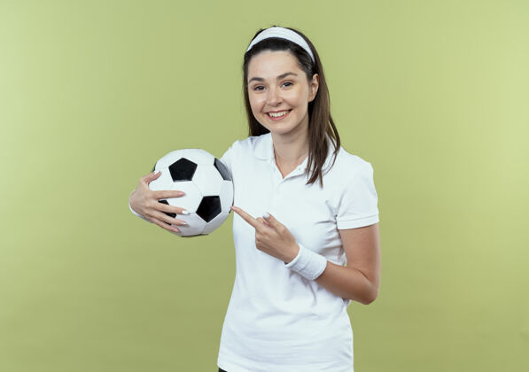 举行戴着头巾的年轻健身女士拿着足球 手指指着足球 站在轻薄的墙上欢快地笑着女子头带健身
