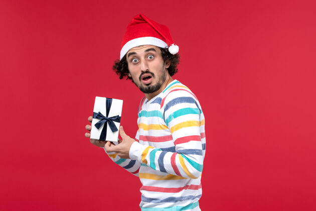 娱乐正面图年轻男性手持新年礼物在红色背景上过年感慨圣诞节人物帽子