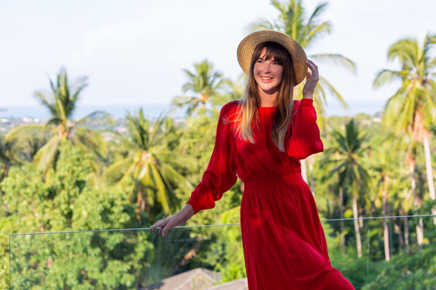 时尚穿着红色夏装 戴着草帽 在阳台上享受热带海景和棕榈树的快乐女人地平线年轻夏季