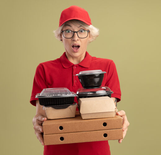 制服年轻的送货员身穿红色制服 戴着帽子 戴着眼镜 手里拿着比萨饼盒和食品包 站在绿色的墙上 惊讶又惊讶年轻人拿着惊喜