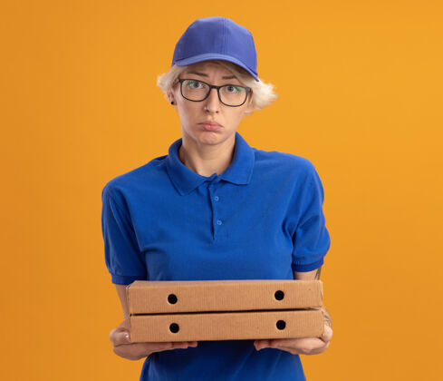 悲伤不满意的年轻送货员身穿蓝色制服 戴着眼镜 拿着披萨盒 表情悲伤地看着橙色的墙上帽子戴着披萨